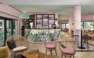 Interior view of the bar and seating area at The Magnolia Hotel featuring pink walls, spotlights, a pastel green pendant light, tables, a wicker style chair, pink chairs, a bar with a white and green leaf design and black wall units behind the bar with bottles of drinks