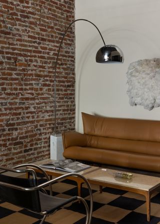 Image of a modern living room with a brown leather sofa and a long and narrow coffee table in front of it. There is a modern black leather and curved metal accent chair across from the sofa. A checkerboard rug is on the ground and there is an arched chrome lamp in the corner.