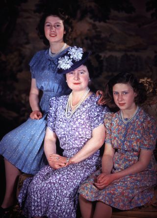 The Queen Mother sitting down wearing a purple dress posing with Princess Margaret and Queen Elizabeth wearing blue floral dresses