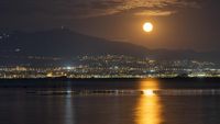 An orange full moon shines over a coastal city at night.