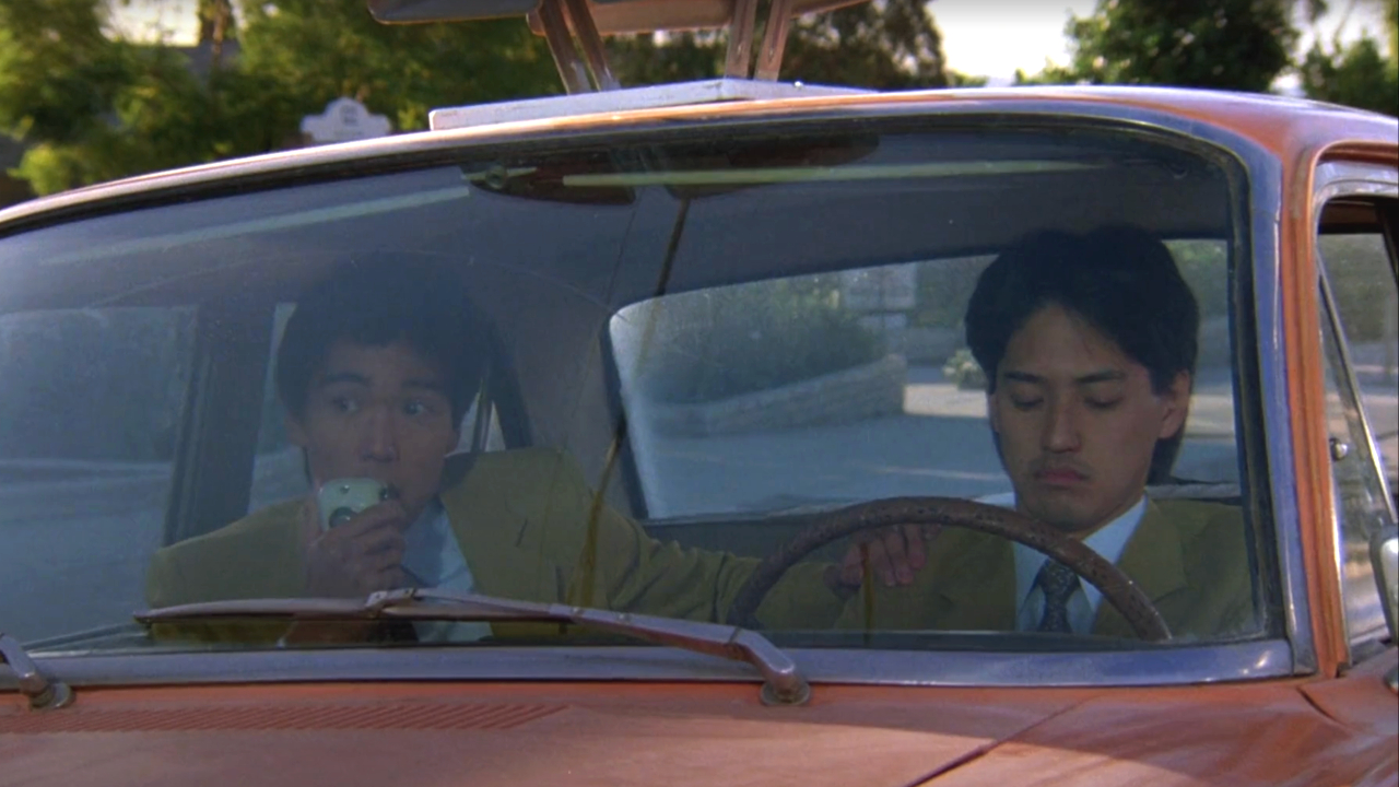 Yuji Okumoto and Brian Imada sit in a car while wearing yellow jackets in Better Off Dead.