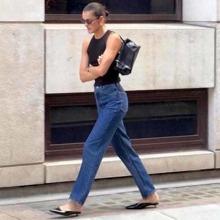 female fashion influencer Valeria @sobalera walks on a London sidewalk with a slick-back bun, oval sunglasses, chunky earrings, black racerback tank top, black shoulder bag, straight-leg jeans, black kitten heel