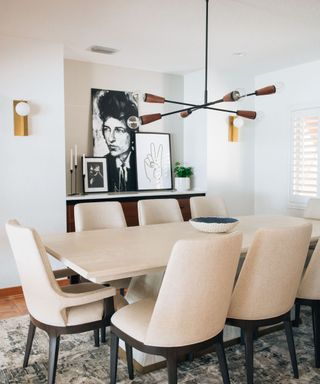 A white mid-century modern dining room with a white wooden dining table with seven upholstered chairs next to it, a brown iron chandelier, and a wooden sideboard with black and white wall art on it