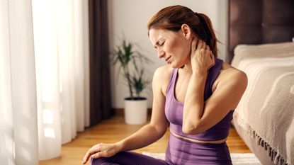 Woman in activewear sits on the floor next to a bed holding the back of her neck and wincing