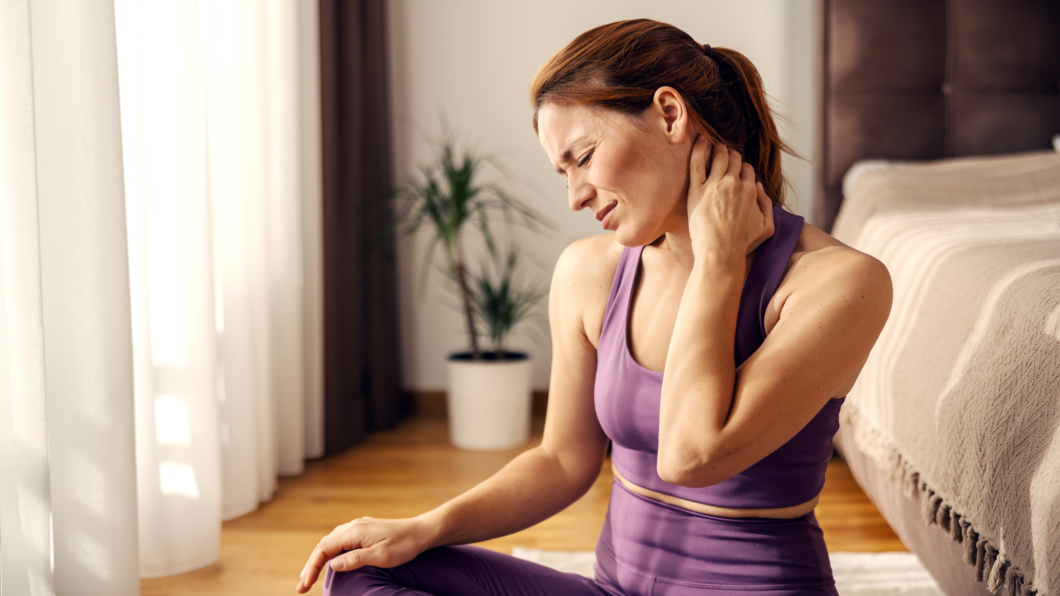Woman in activewear sits on the floor next to a bed holding the back of her neck and wincing