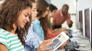 Teens work on desktop and tablet computers while teacher answers a question.
