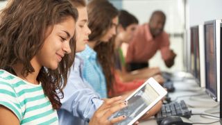 Kids work on desktop and tablet computers while teacher answers a question.