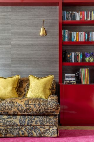 media room/library with red gloss bookcases, patterned couch with gold damask cushions, gold wall light, pink rug, gray textured wallpaper