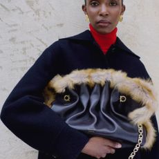 Woman wearing navy coat, red turtle neck, faux leather and fur bag, gold earrings, and staring directly at camera.