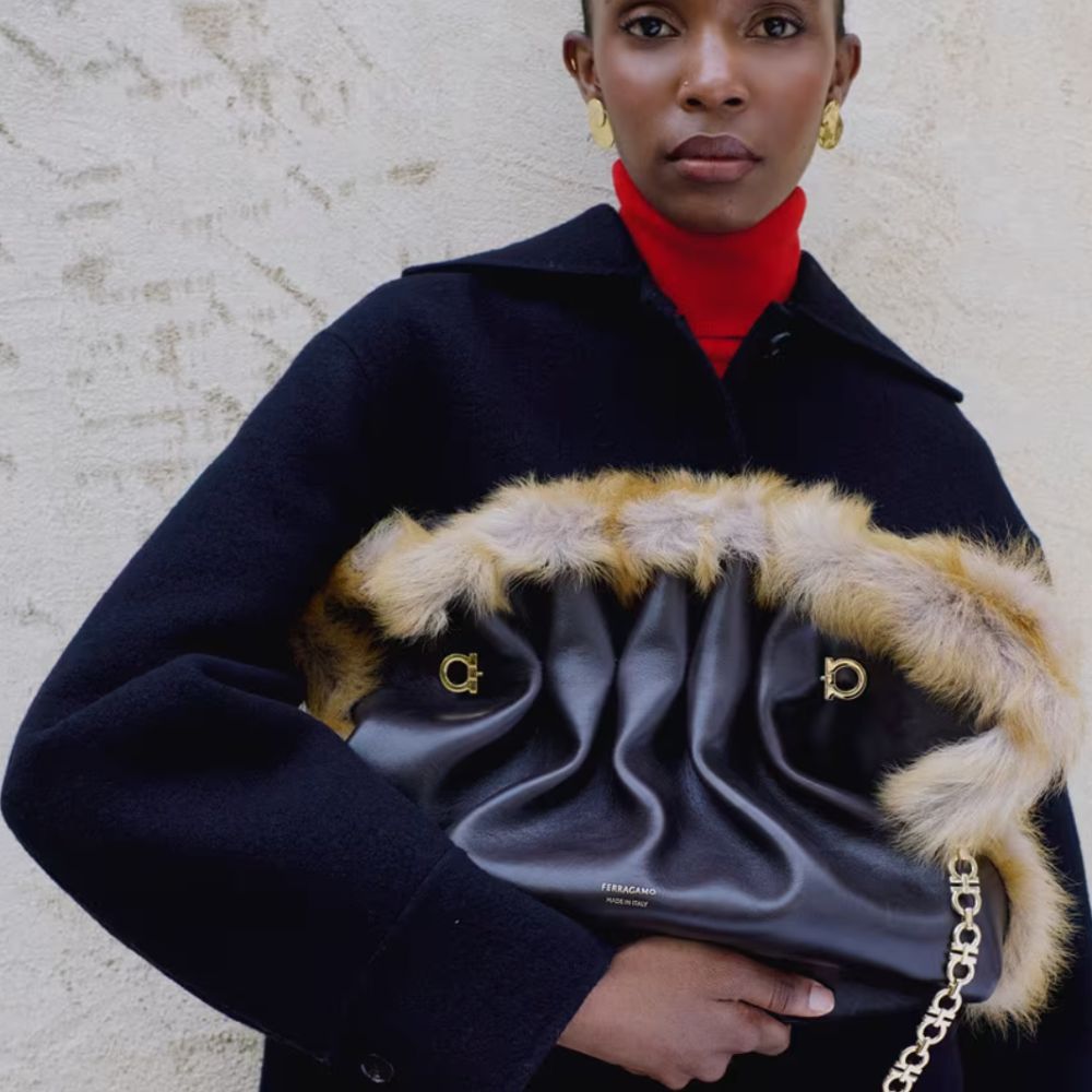 Woman wearing navy coat, red turtle neck, faux leather and fur bag, gold earrings, and staring directly at camera.