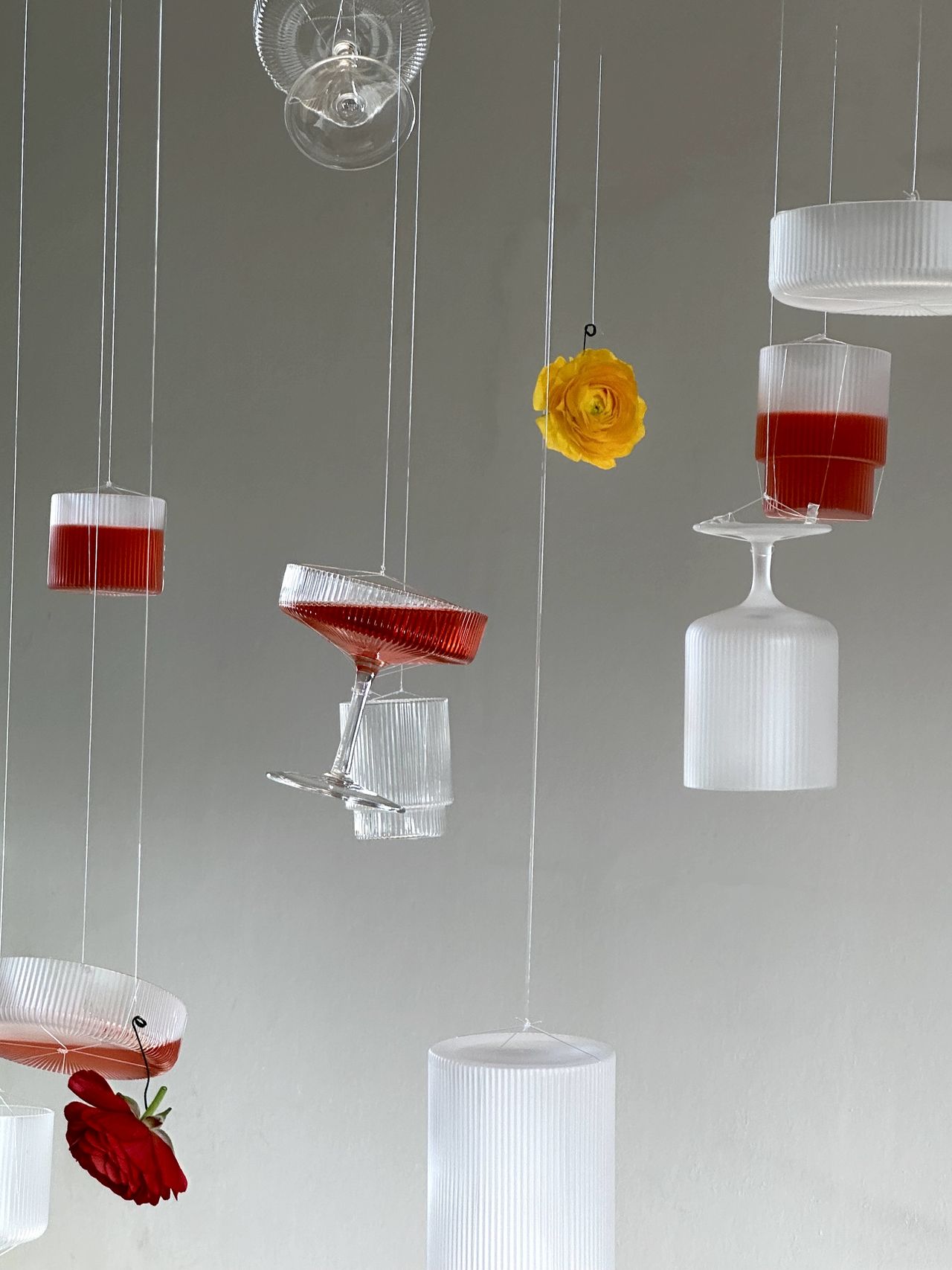 A series of glasses hangs from the ceiling in a white room, filled with red liquid and coupled with flowers.