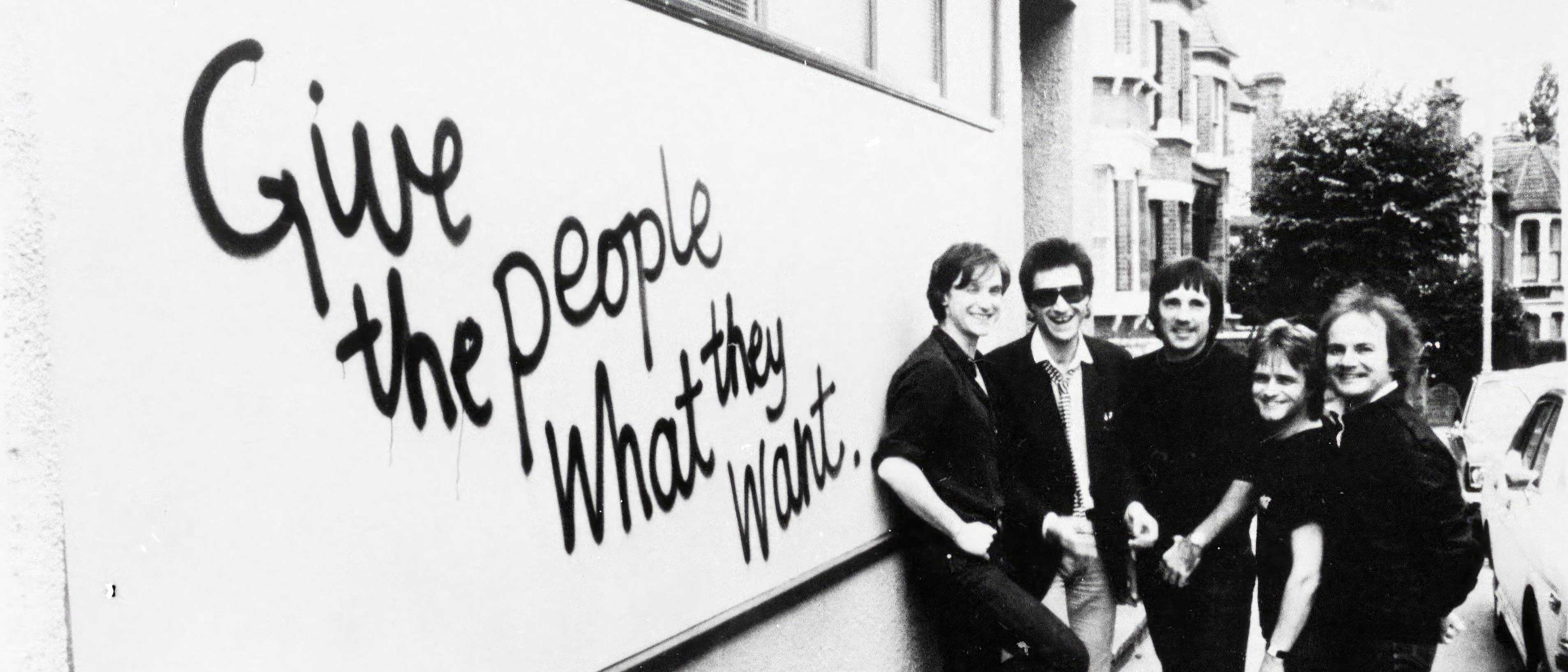 The Kinks standing next to a Give The People What They Want billboard