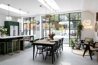 Industrial-style kitchen-diner with concrete floor, green kitchen and island, black wooden table and chairs, and seating area with rattan lounge chairs and 'let's stay home' neon light