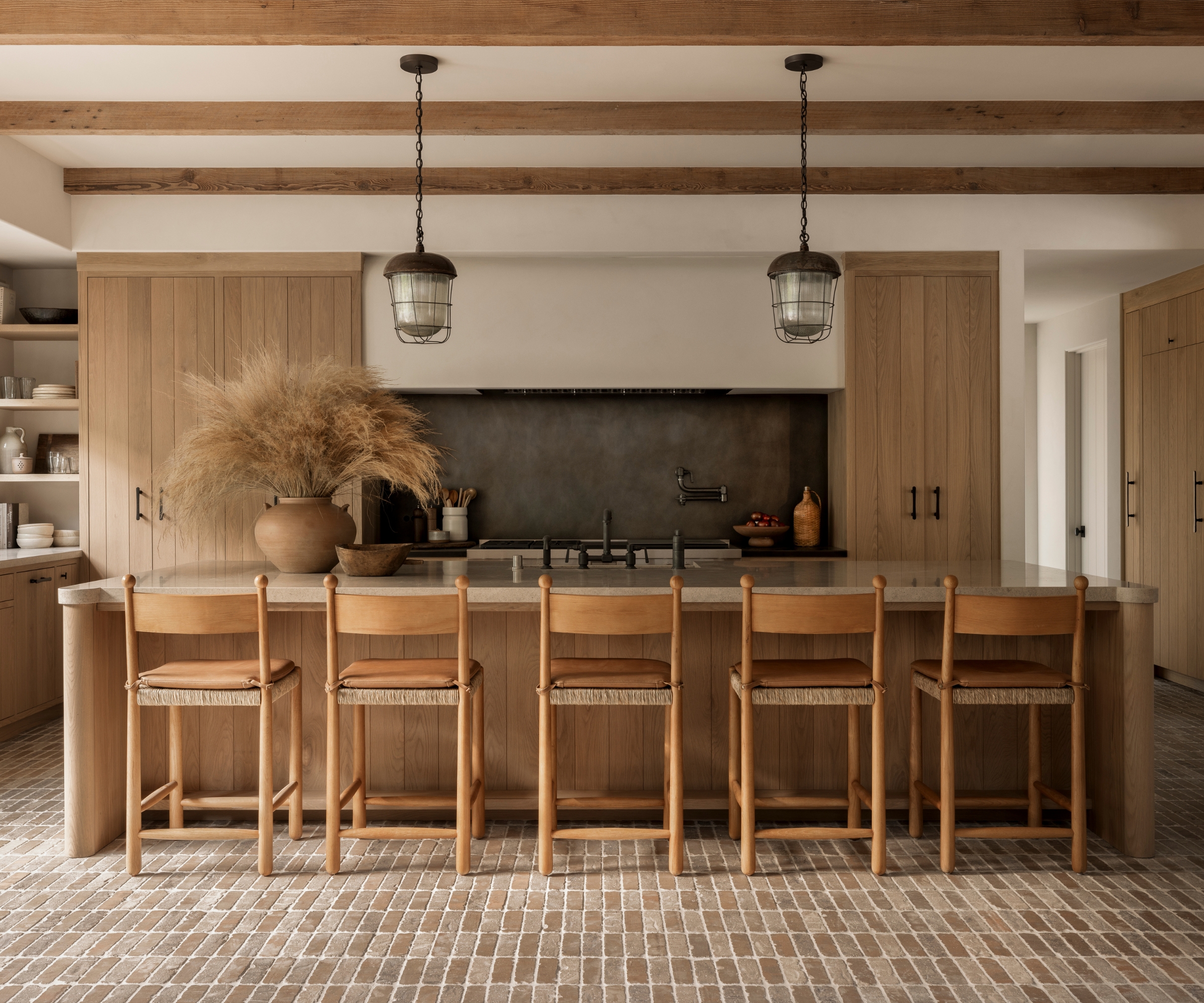 A wooden kitchen with shiplap cabinet doors, a black patinated metal backsplash, brick floors and exposed ceiling beams