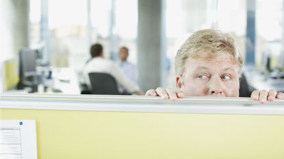 A man looking fearful looks over the top of a cubicle wall in an office.