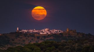 a large orange moon hovers over a distant castle built wide across the top of a hill or small mountain.