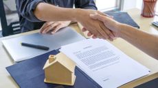 Close-up of two hands shaking over a desk with a contract to buy a home on top of it, and a wooden miniature house model 