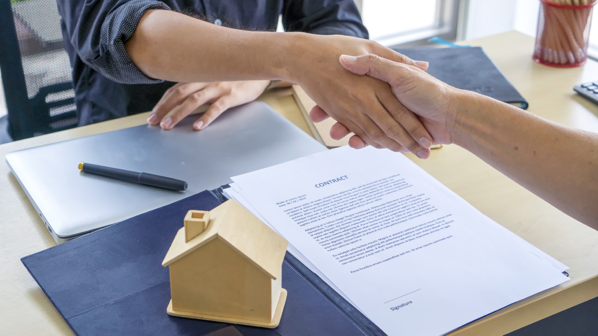 Close-up of two hands shaking over a desk with a contract to buy a home on top of it, and a wooden miniature house model 