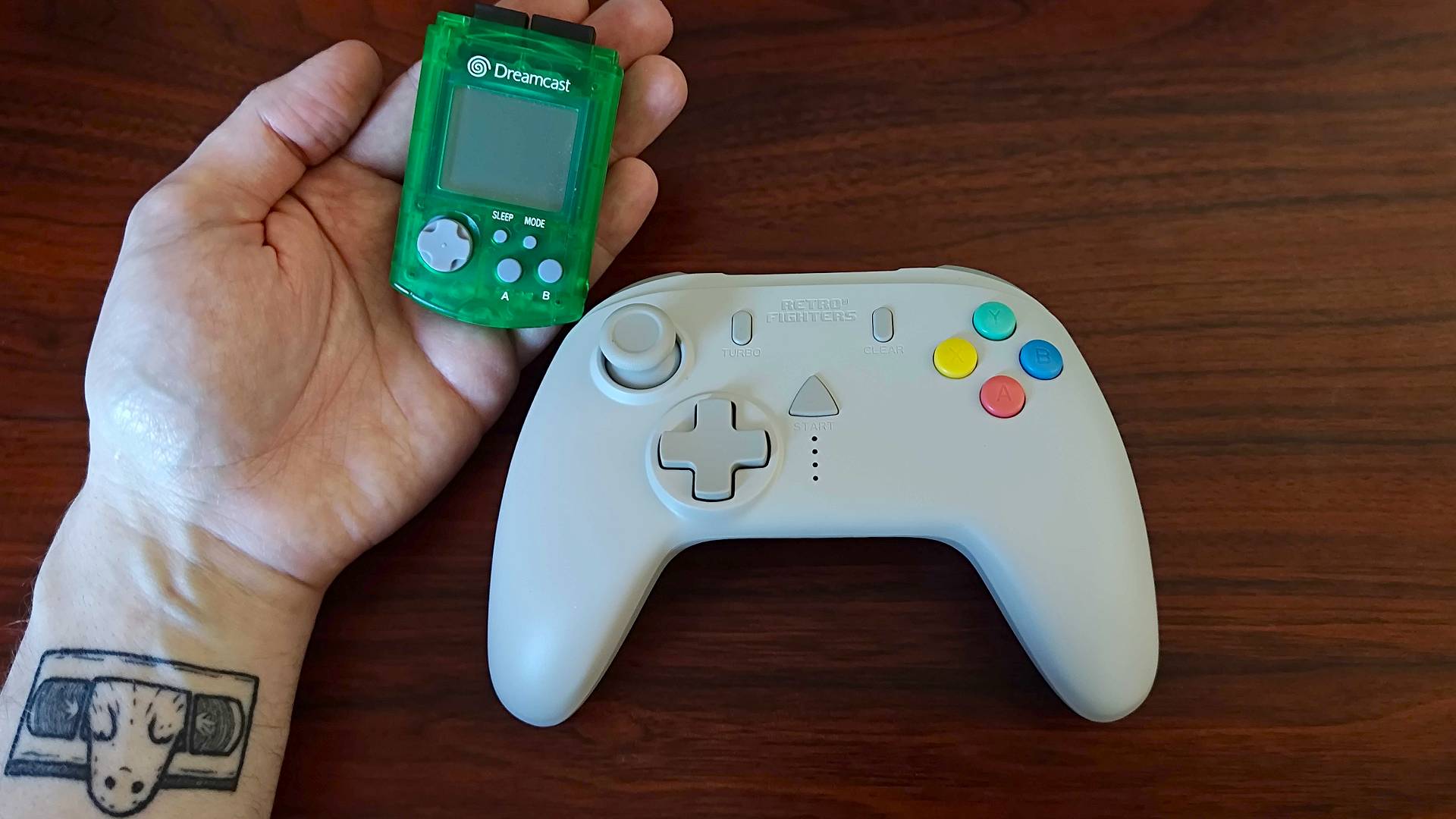 Hand holding green Dreamcast VMU next to Retro Fighters StrikerDC controller on woodgrain desk.