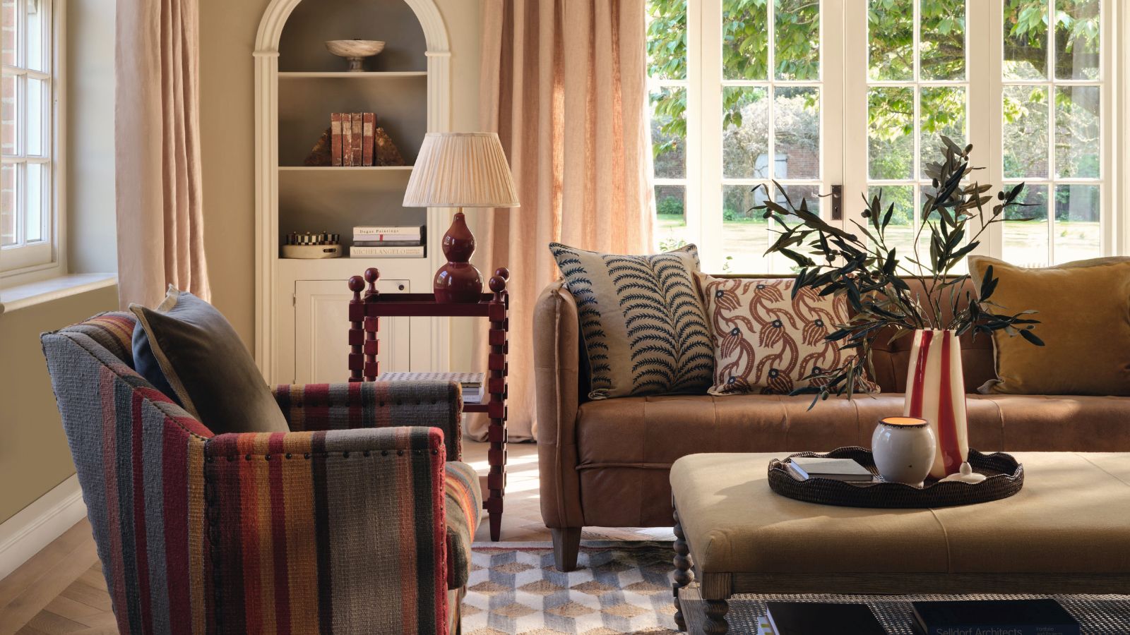 A warm-toned living room, with a brown three seater sofa and a striped arm chair. An upholstered ottoman table between them, and large glass patio doors behind. 