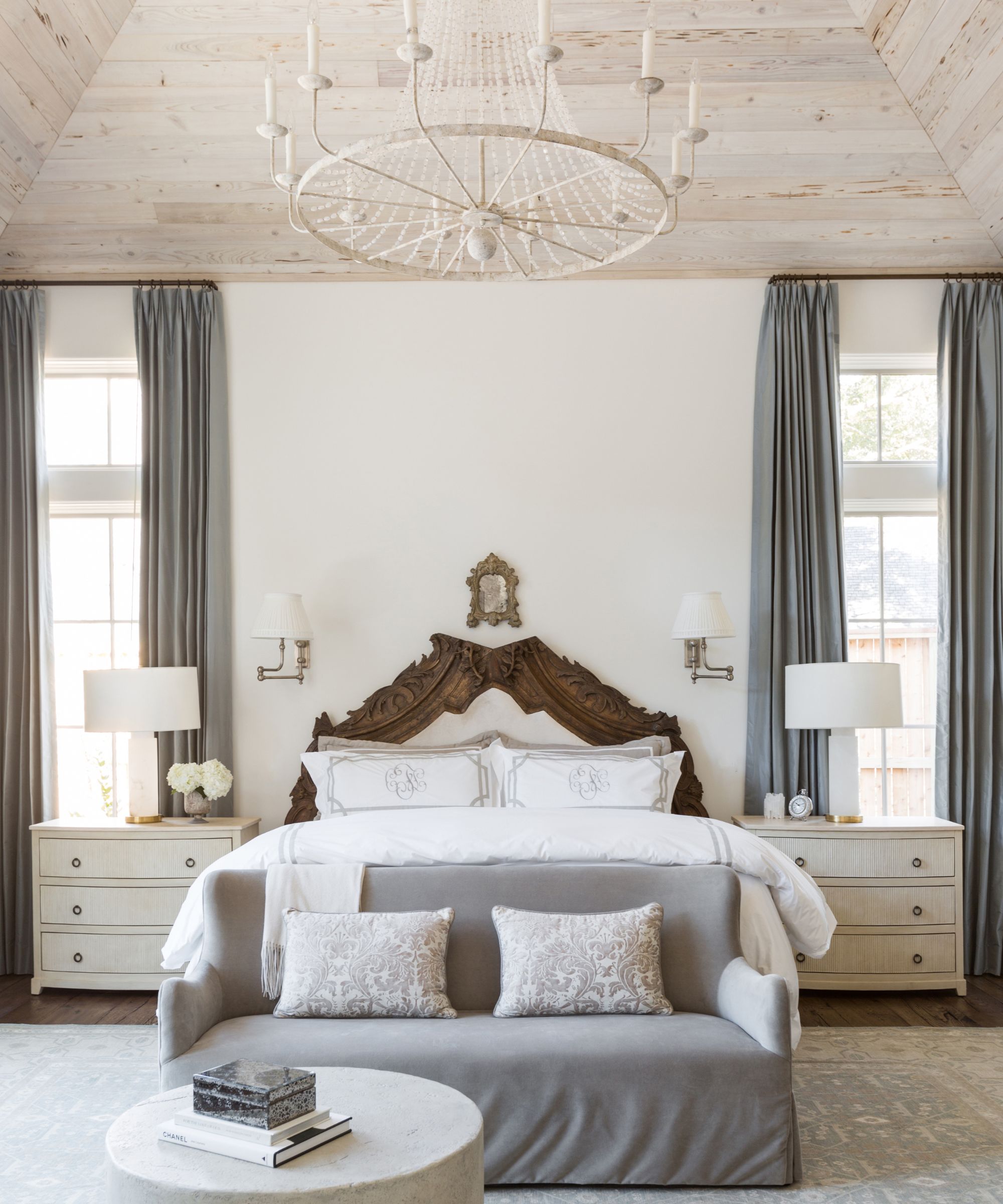 Bedroom with exposed bleached wood ceiling, a large white chandelier, tall, long windows with grey drapes, an antique wood carved bed with white monogrammed bedding, and a grey velvet sofa at the end