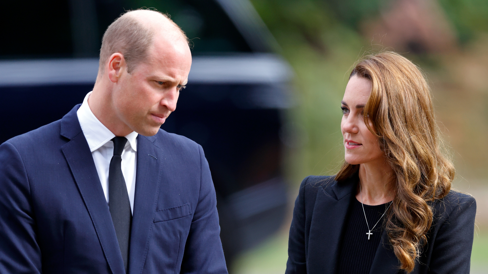 Prince William wears a navy suit and looks down at the ground, while wife Kate Middleton wears a black suit and has long brown golden hair