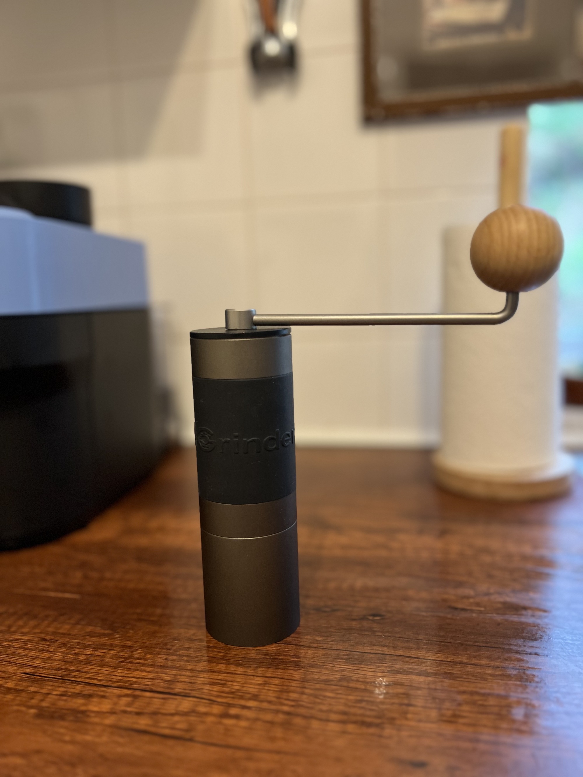 A gray and black manual coffee grinder with a handle that has a wooden ball on the end. The grinder is on a wooden countertop.