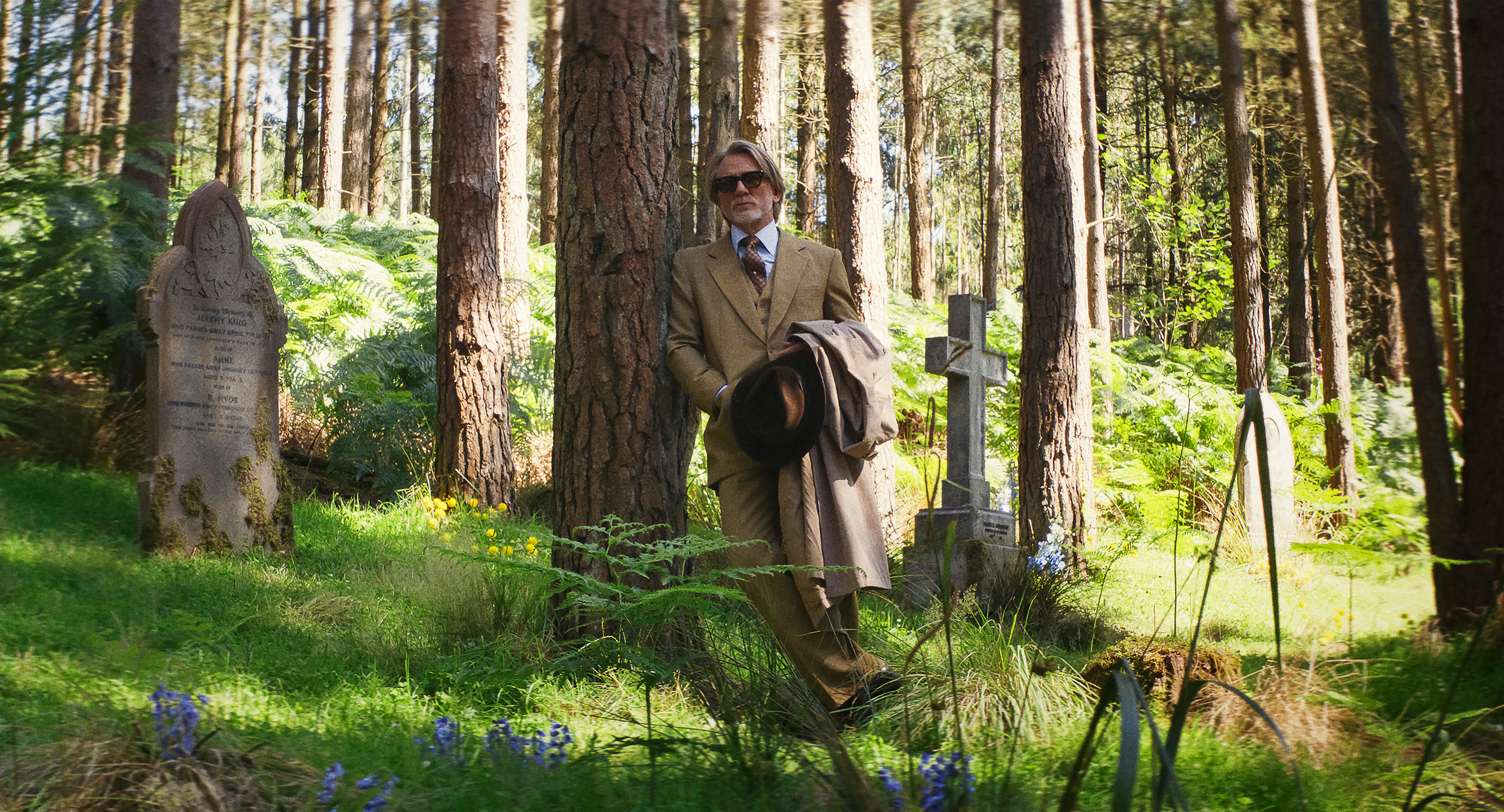 benoit blanc wearing a tan suit leaning against a tree in a cemetery in wake up dead man