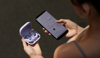 A woman holding a set of earbuds reported to be the Beats Powerbeats Pro 2, also holding a smartphone with the companion app, about to toggle 'Heart Rate'