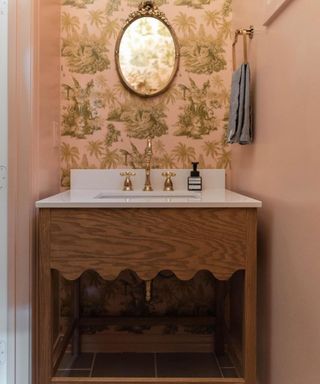 A pink bathroom with pink and green wallpaper, a vintage mirror, a scalloped sink, and a towel hook with blue towel
