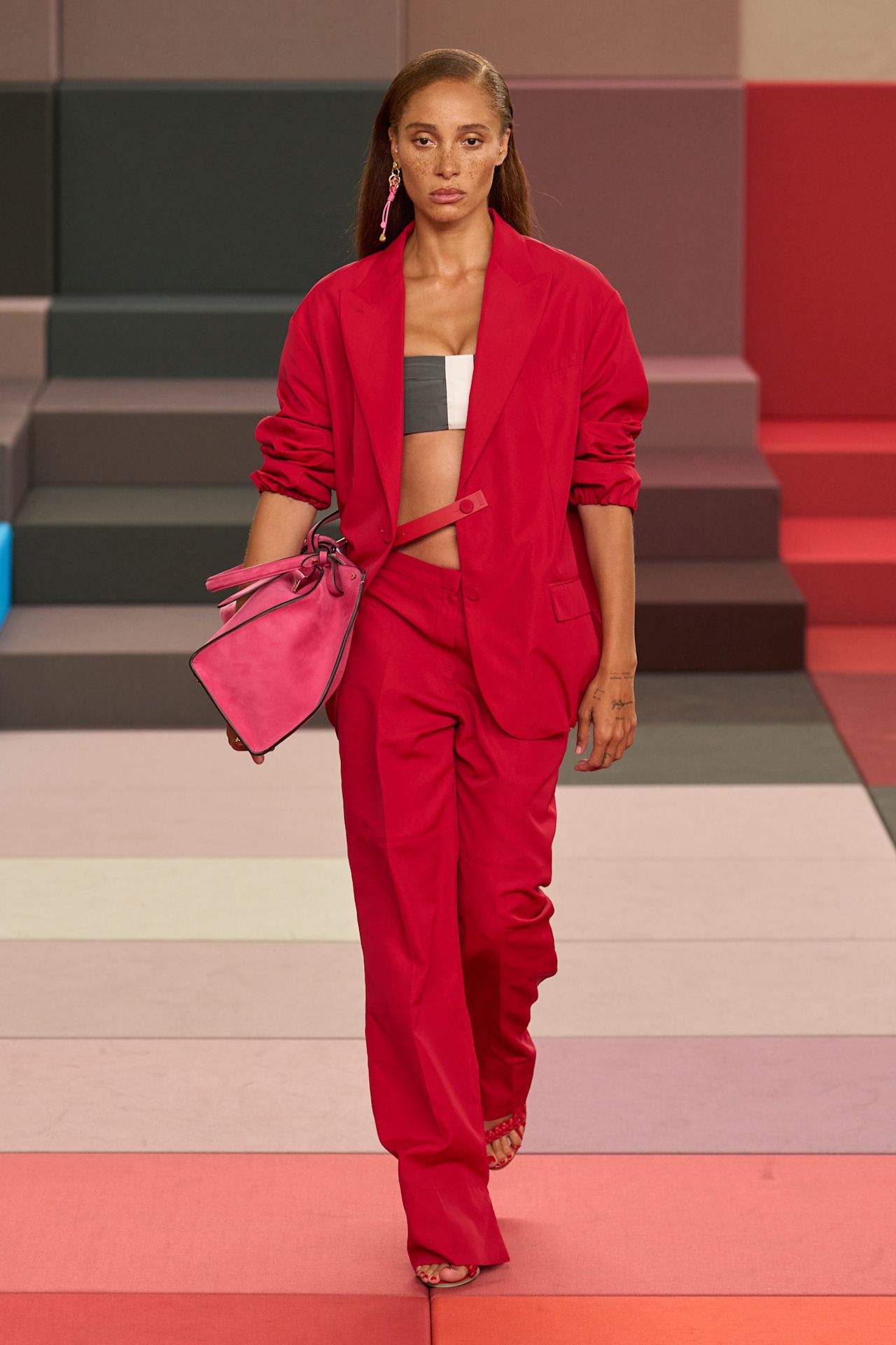 A Fendi model wearing an oversize red blazer worn over a two-tone gray and white bra top, matching bright red pleated wide-leg pants, red sandals, a bright pink suede handbag, and a single pink chain asymmetrical earring.