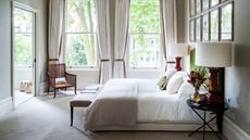 A bright and white carpeted minimalist bedroom with a large bed with white and beige bedding and green headboard, black circular bedside tables, red lamps with white lampshades, a framed gallery wall and a brown wicker chair. 