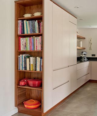 A kitchen with light pink cabinets and an open wooden shelf with colorful books and red and orange dutch ovens on it