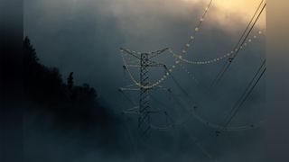 A tall power line tower stands against a dramatic, cloudy backdrop, with multiple power lines extending from it. The lines are adorned with illuminated insulators, adding a subtle glow to the scene. Dark silhouettes of trees are visible on the left side, contrasting with the lighter sky above.