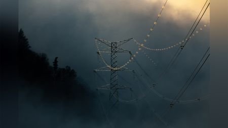 A tall power line tower stands against a dramatic, cloudy backdrop, with multiple power lines extending from it. The lines are adorned with illuminated insulators, adding a subtle glow to the scene. Dark silhouettes of trees are visible on the left side, contrasting with the lighter sky above.