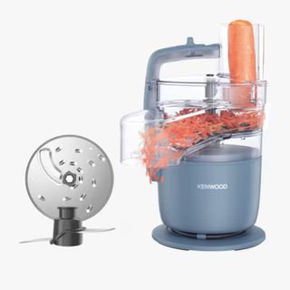 A compact countertop food processor shredding a carrot with an additional blade pictured to the left