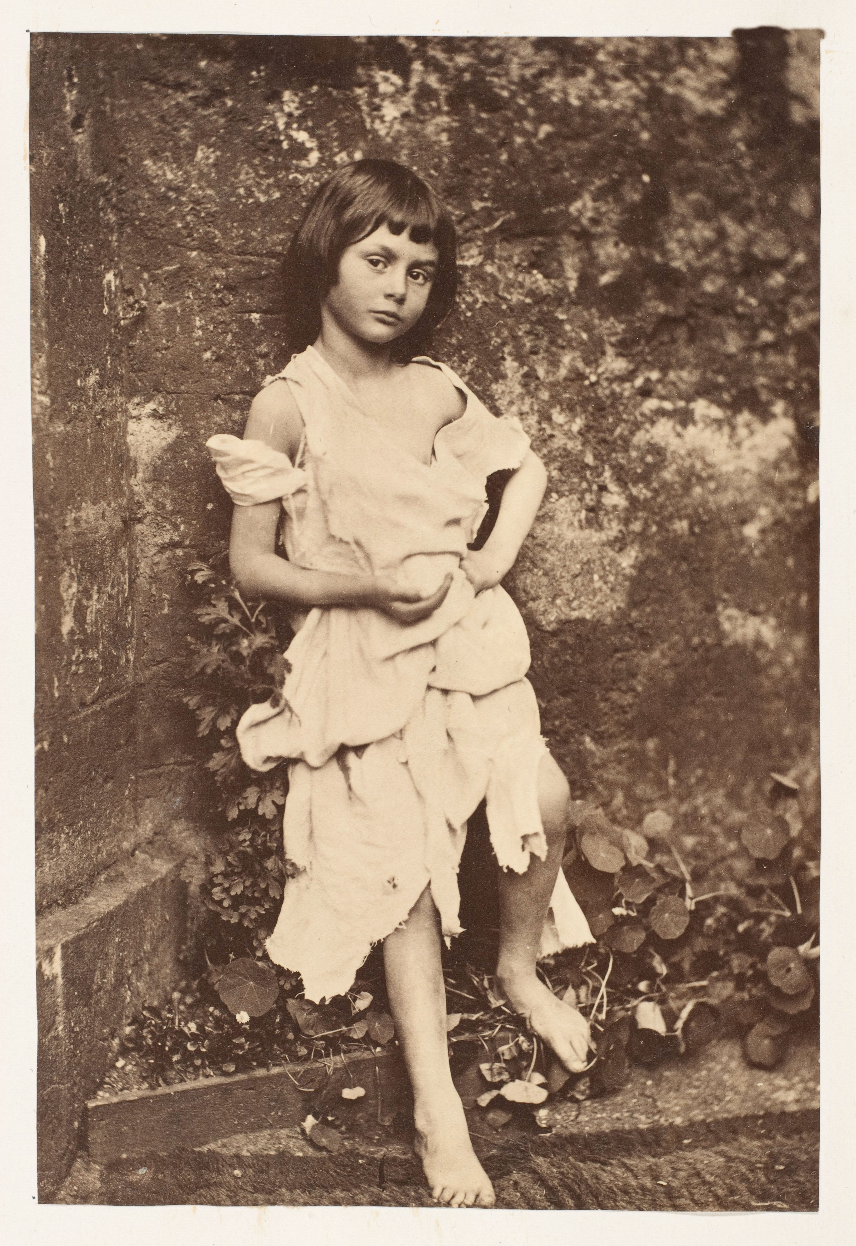A vintage sepia-toned portrait of a young, barefoot girl with short dark hair wearing a loose, tattered white dress while leaning against a textured stone wall.