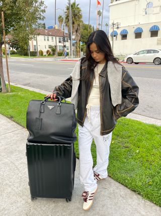 a woman standing on the sidewalk with a suitcase and tote bag wearing a leather jacket and sweatpants