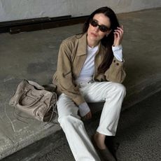 Woman wears suede jacket, white long sleeved top, white jeans and carries taupe balenciaga city bag