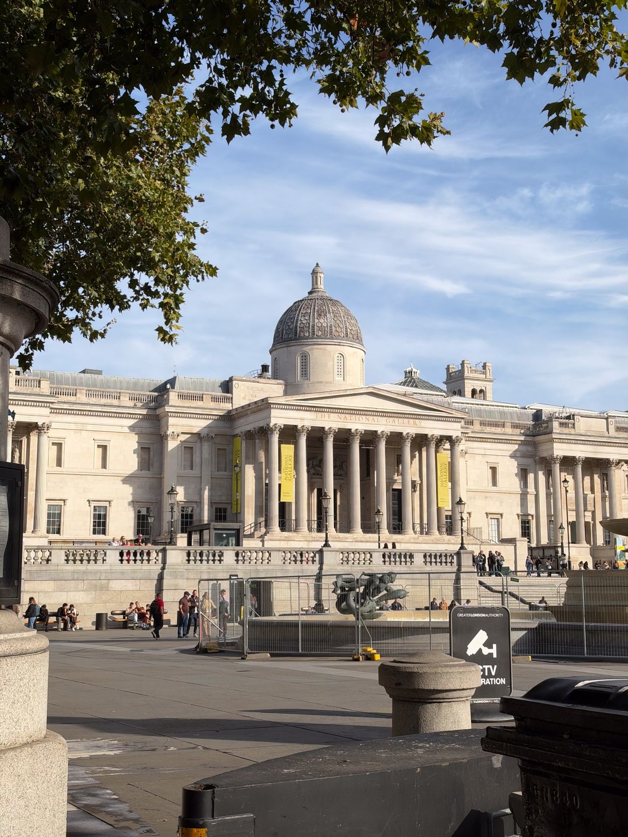 National Gallery in Trafalgar Square framed by leafy tree branches on a sunny day.