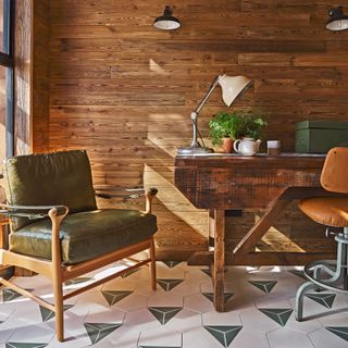 A modern traditional home office garden room with black light fixtures, wood wall panel decor, olive green leather accent chair with wooden legs, tan leather desk chair and off-white and green floor tiles with triangular motif