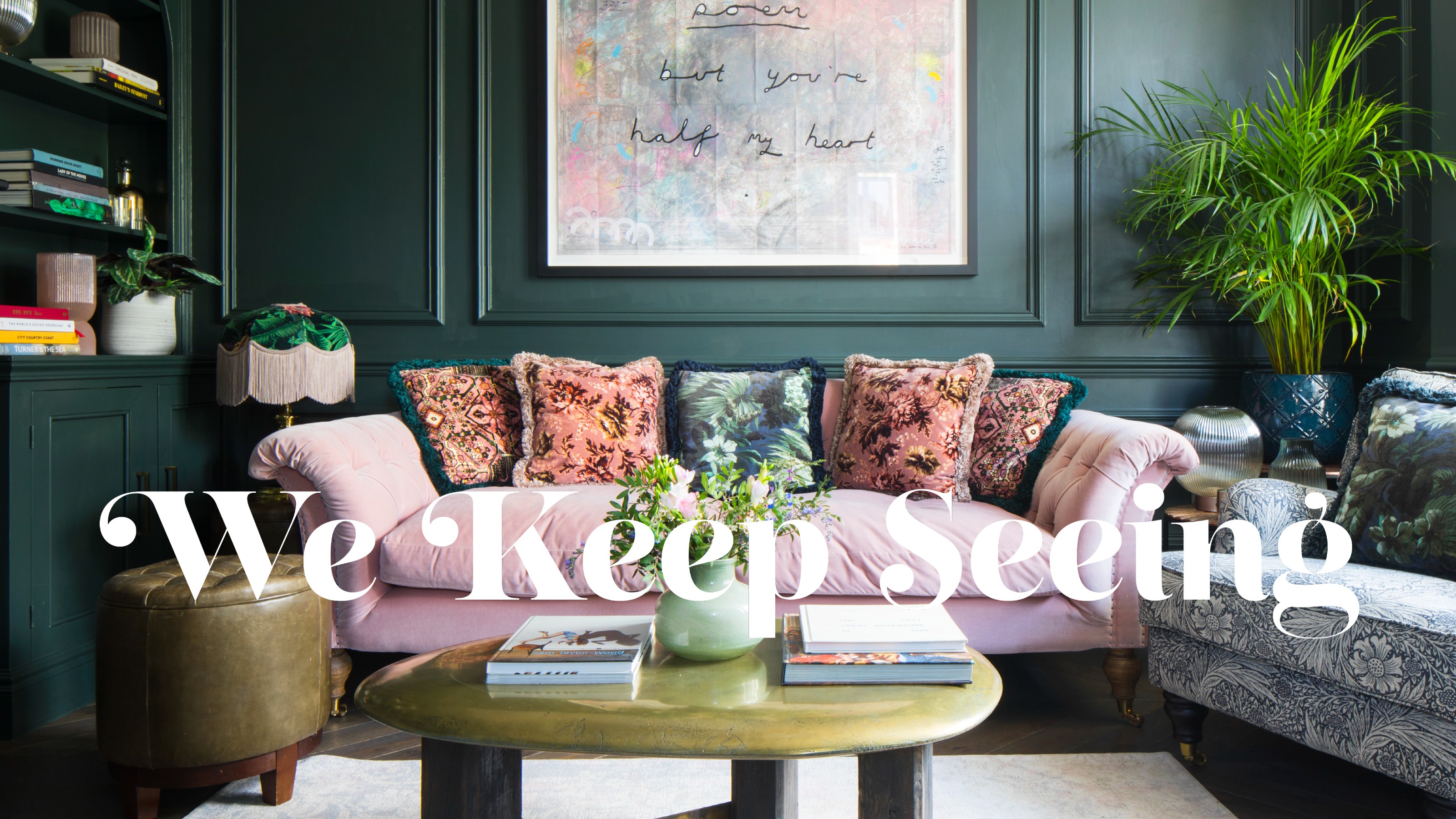 A dark green-painted living room with a pink velvet sofa with patterned, fringe-trimmed cushions on top and a fringed table lamp next to it