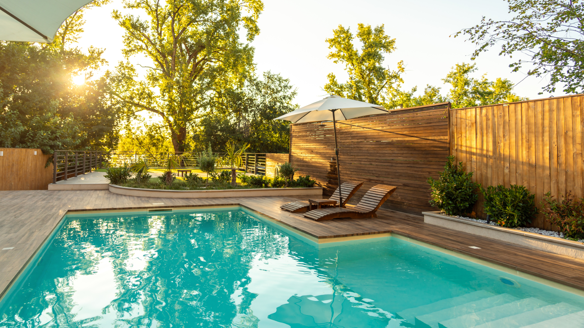 outdoor pool with wooden decking, wooden fence, planted areas and two wooden sun loungers and patio umbrella