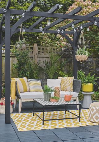 A patio and pergola with an outdoor sofa, coffee table sat on a yellow rug, and string lights