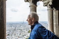 A smiling retired man traveling in Europe. He is framed by stone arches, viewing the cityscape and the Eiffel Tower from Sacre Coeur in Paris, France.
