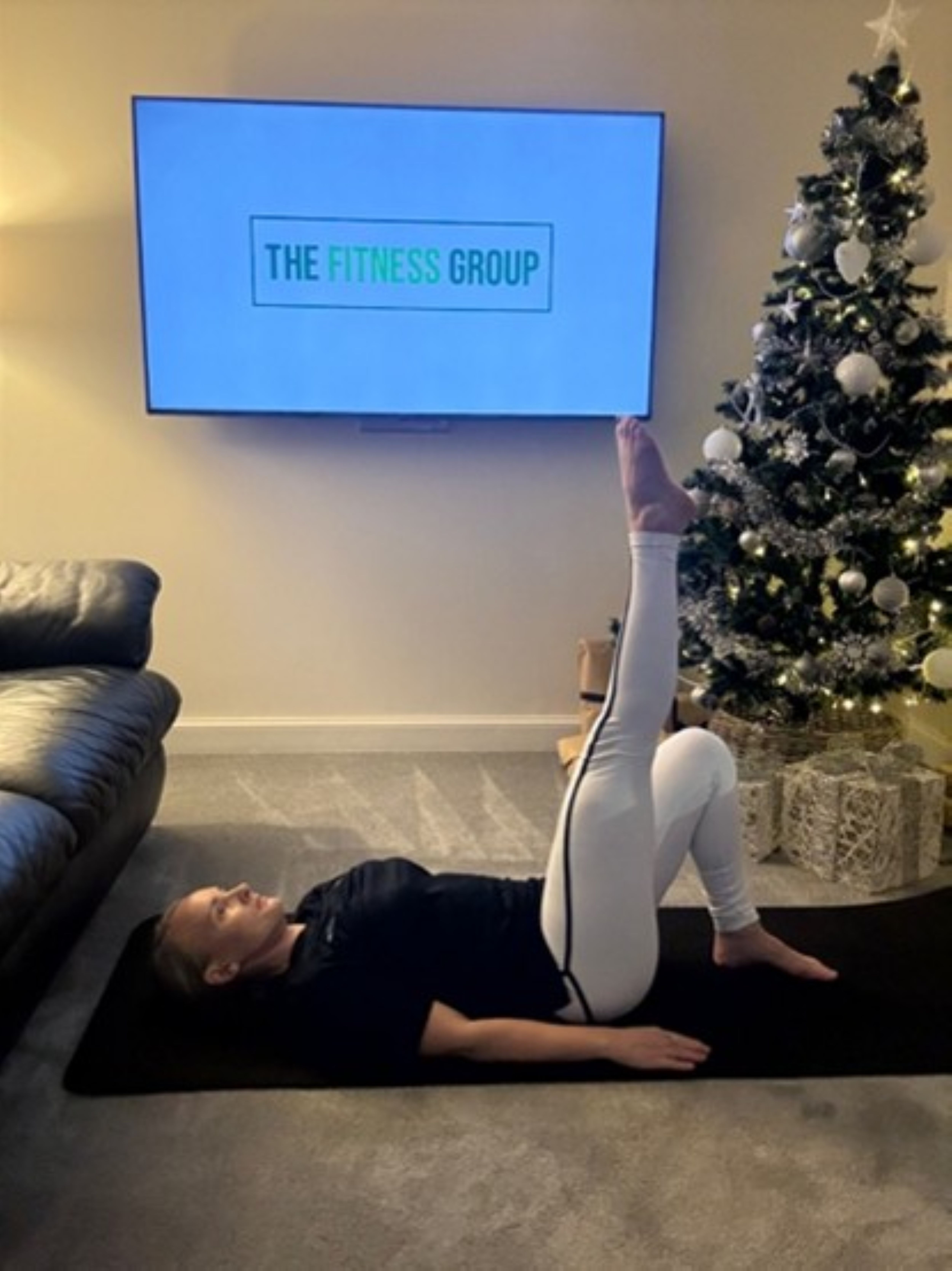 Woman demonstrates a Pilates exercise between a sofa and a Christmas tree. A mounted TV in the background reads "The Fitness Group"