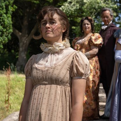 mary bennet wears a tan dress and glasses and stares off in the distance as her family watches on in a still from the other bennet sister