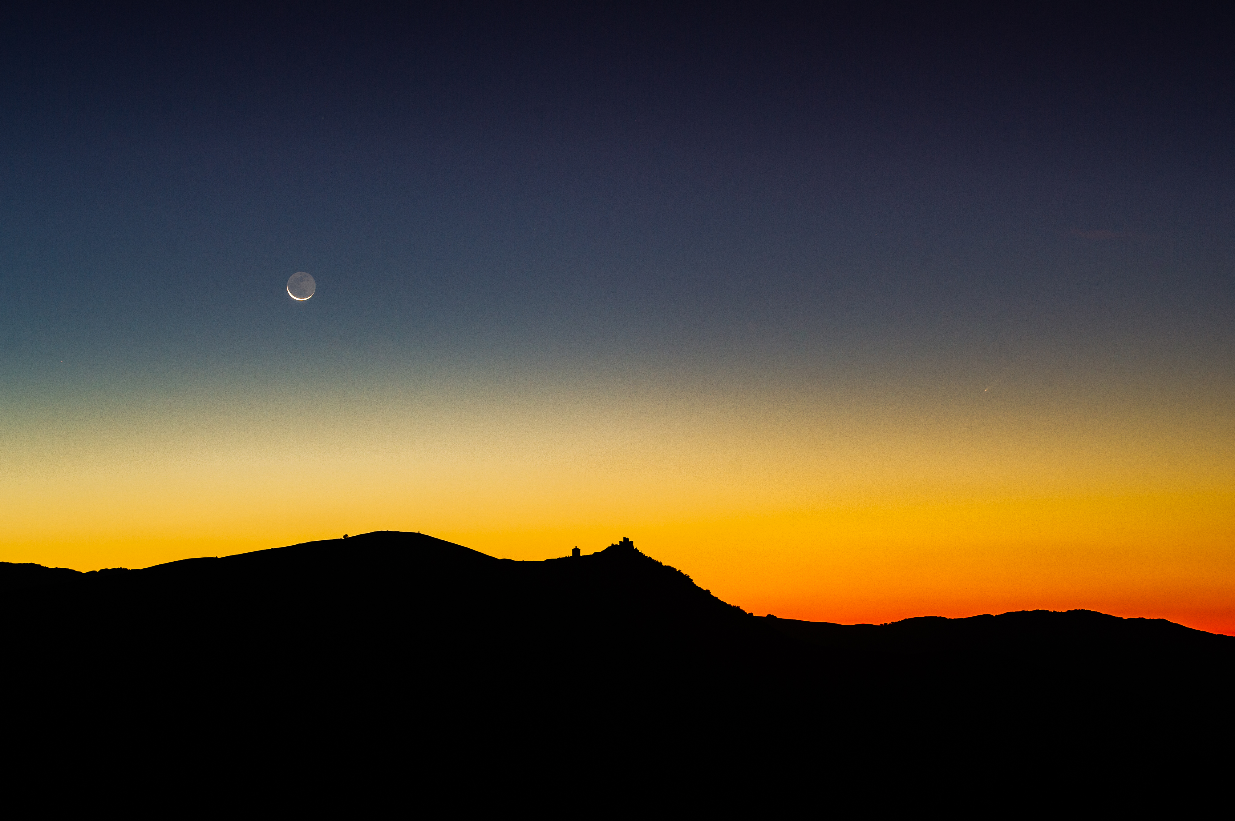 a silhouetted lanscape below with a small church and in the sky a thin waning crescent moon with a faint fuzzy comet on the right. They sky is painted yellow and orange as the sun prepared to rise.