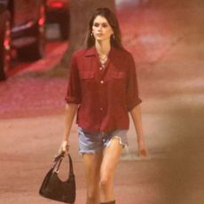 Kaia Gerber walks down the street wearing a red boxy shirt, denim cut-off shorts and knee-high boots. She carries a black bag and accessorised with necklaces and hoop earrings.