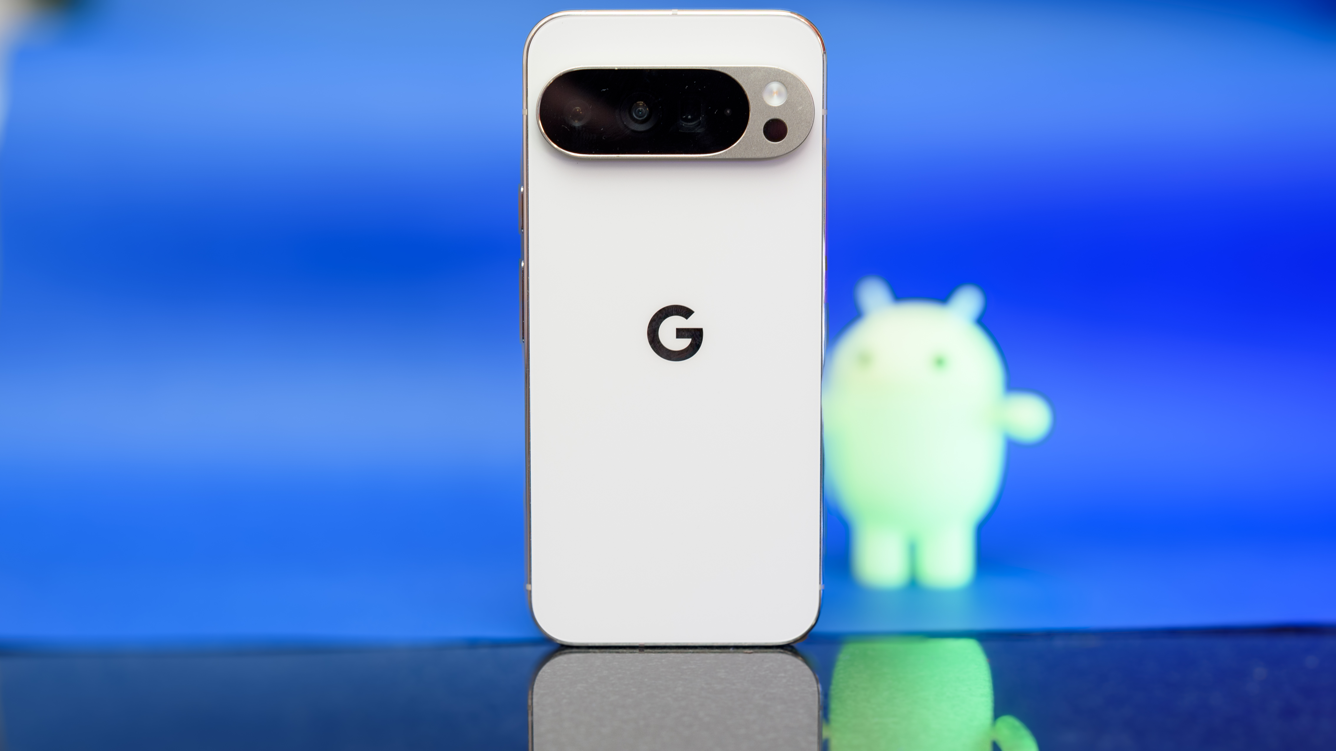 Google Pixel 10 Pro XL in Porcelain against a blue background with a green neon Android figurine in the background